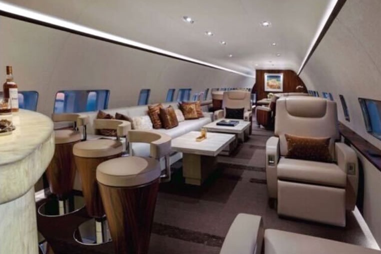 The interior of a plane, beige reclining seats and bar stools can be seen