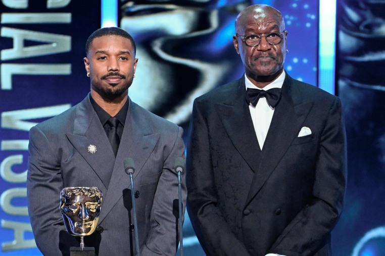 Michael B. Jordan and Delroy Lindo stand at a lectern on stage during an award ceremony. They look nonplussed.