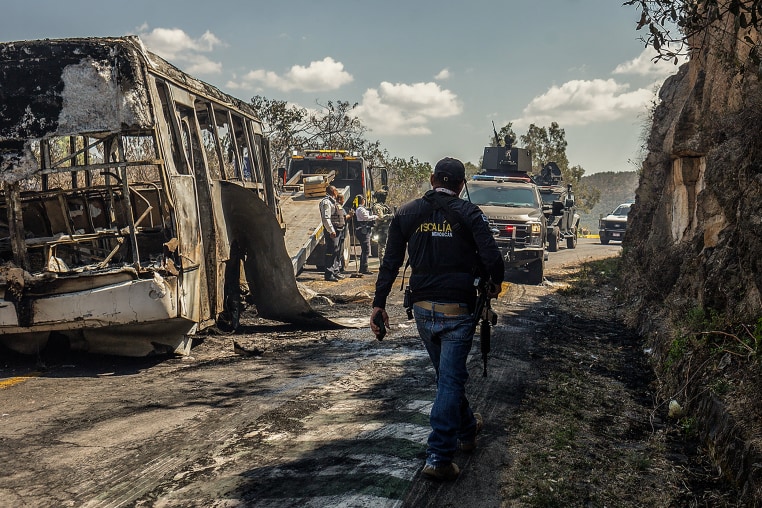 A charred bus sits in the middle of a mountain road. Law enforcement officers stand nearby.