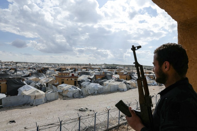 A person holding a gun looks towards a village of tents outside