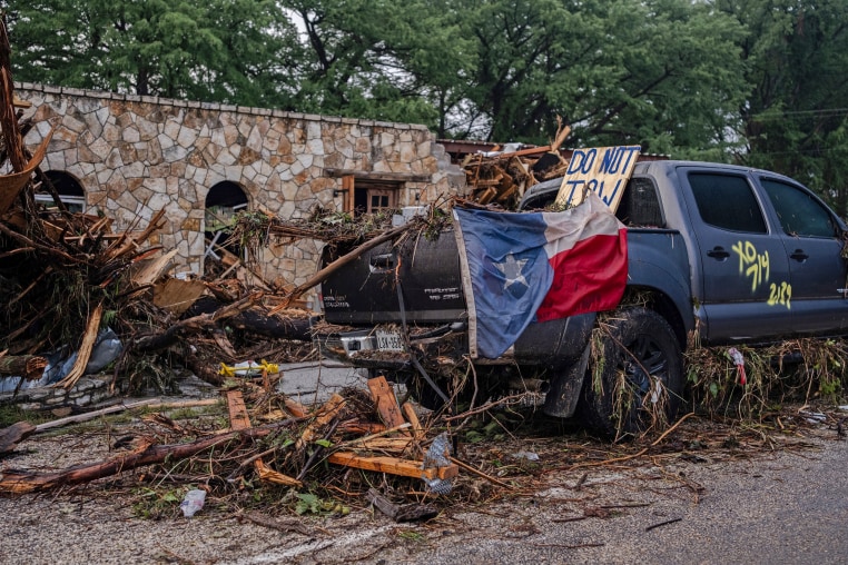 A destroyed SUV draped in a Texas State flag sits next to the road in debris.