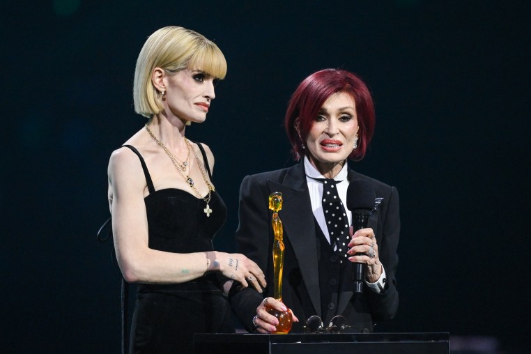 Kelly Osbourne, left, and Sharon Osbourne stand at a podium on stage, while Sharon holds an award statue and speaks