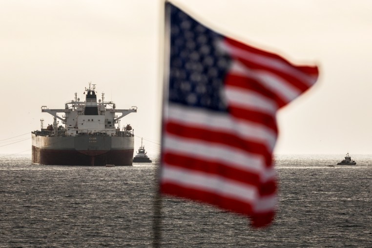 An American flag appears in soft-focus in the foreground. Behind it several ships float in the water.