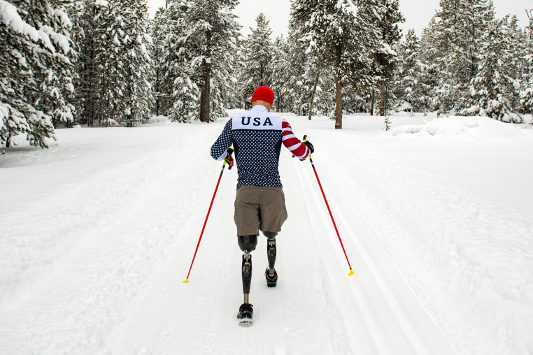 A man with two prosthetic legs  walks in a snowy landscape using two red hiking poles.