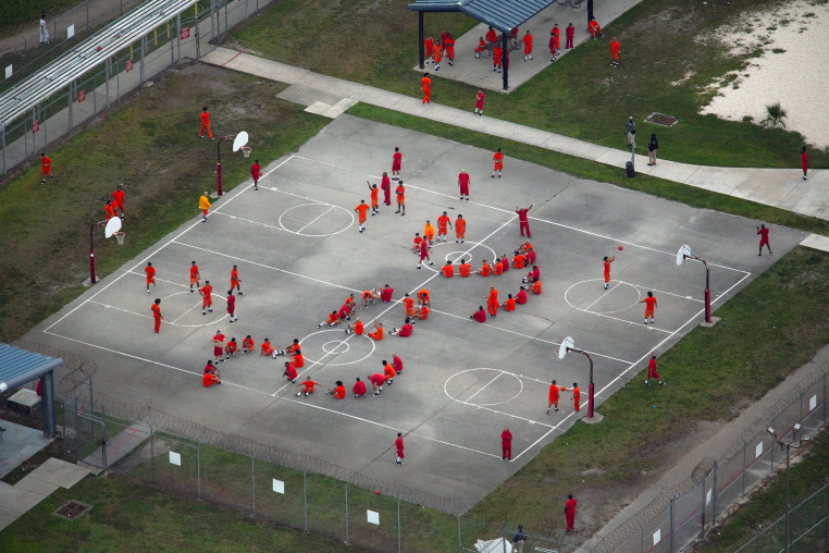 An aerial view of dozens of detainees wearing orange prison uniforms on a basketball court, arranging their bodies to spell SOS.