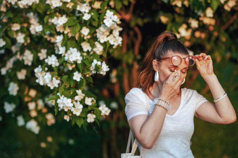 Summer time. unhappy elegant female in white shirt with handkerchief and eyeglasses has an allergy attack near flowering tree.