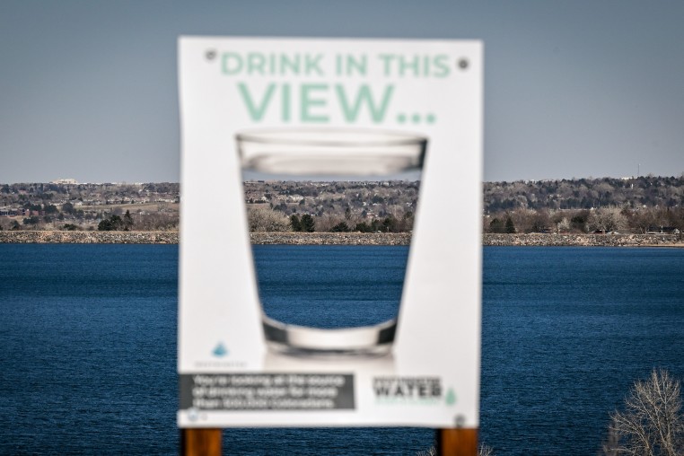 A sign with a water glass on it appears in soft focus in front of a large mountain lake. 