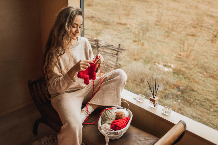 Young Woman Holding Needles and Knitting Handmade Piece From Red Yarn while Sitting near Window.