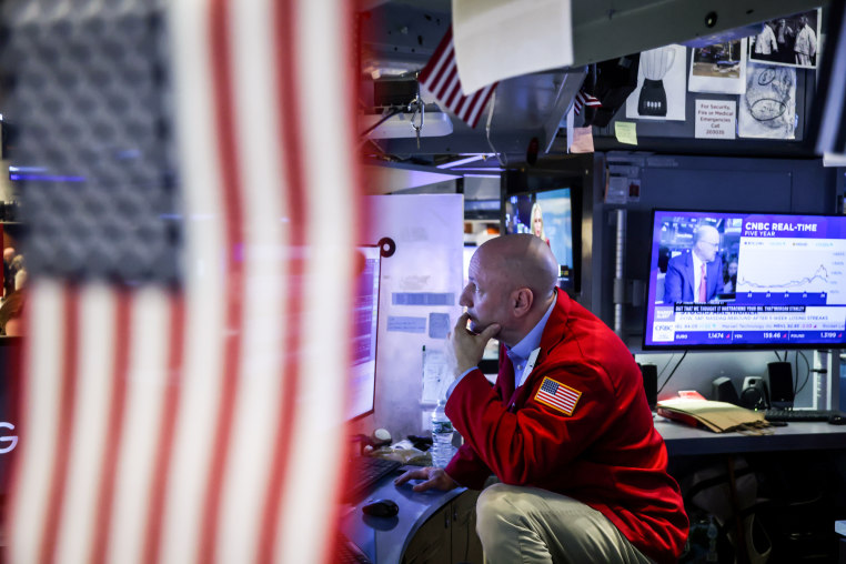 A person sits at a work desk with various monitors, an American flag covers half of the photo in the foreground