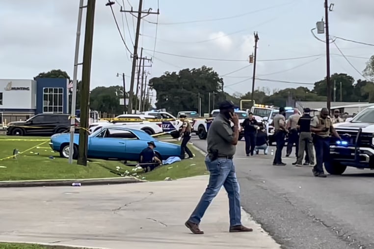 Yellow police tape stretches across a street where numerous law enforcement officials examine a crash site.