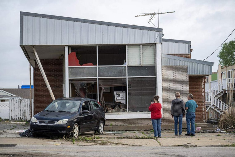 People stand outside of a damaged building with a damaged car parked out front