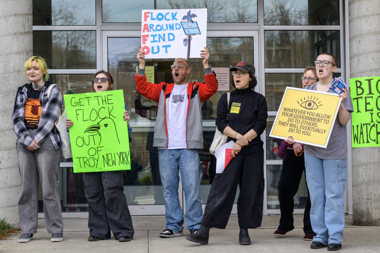 Six people hold signs in front of a glass doorway during a protest against surveillance cameras.