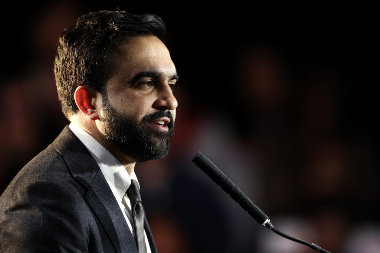 Zohran Mamdani speaks into a small microphone against a blurred background.