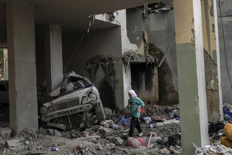 A young girl wearing a white headscarf walks through the rubble of a destroyed concrete building.