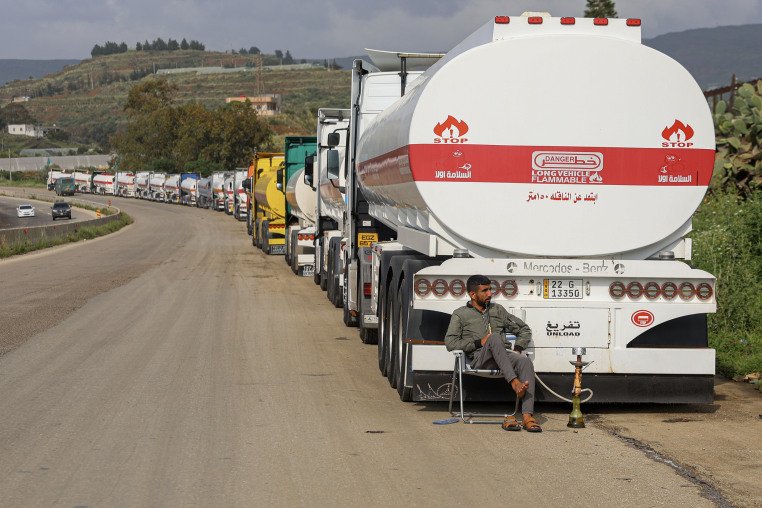 A man sits on a folding chair in front of a long line of tanker trucks parked alongside a dusty highway. 