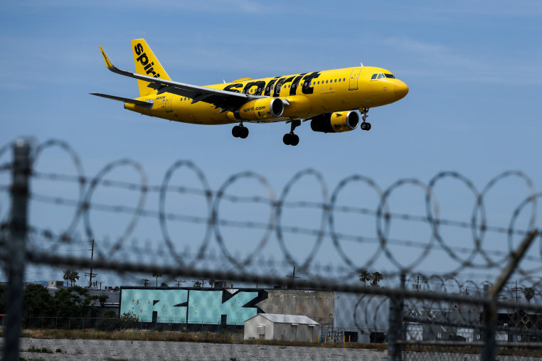 A yellow Spirit Airlines planes can be seen flying above a barbed wire fence in the foreground