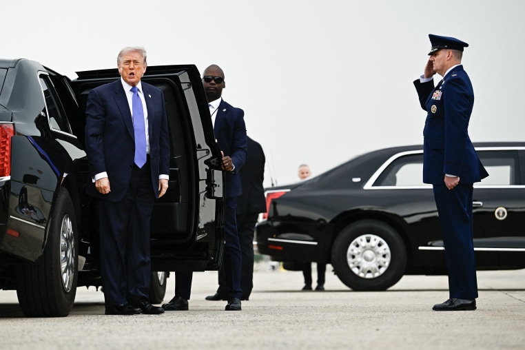 Donald Trump stands in front of a black limousine on a tarmac with his mouth open while speaking.