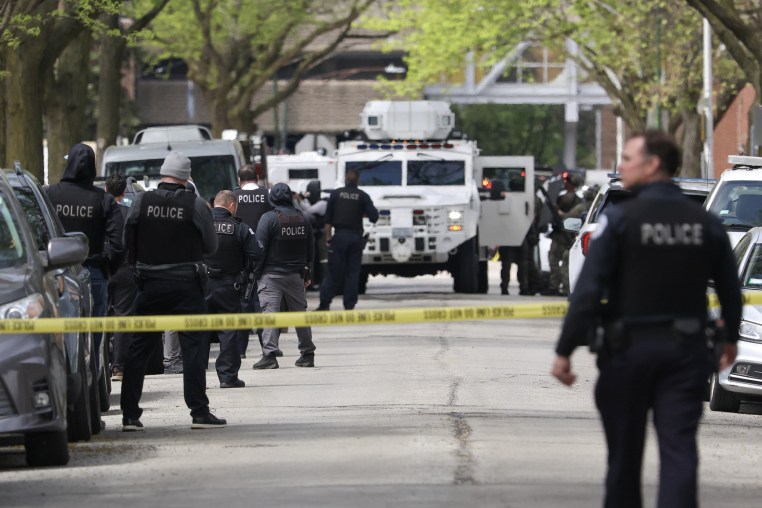 Several police officers in black flak jackets stand behind yellow tape in a city street.