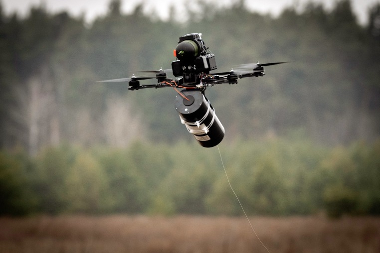 A wired drone hovering in front of a distant treeline.