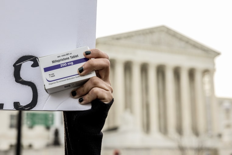 A hand with black fingernails holding up a box of Mifepristone abortion pills in front of the U.S. Supreme Court building.