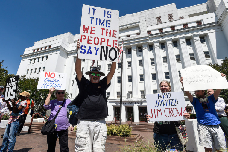 People hold signs outside against redistricting, slogans such as "It's About Us" / "No to Election Rigging" / "It is time we the people say no" / and "Who invited Jim Crow?" can be seen written on signage