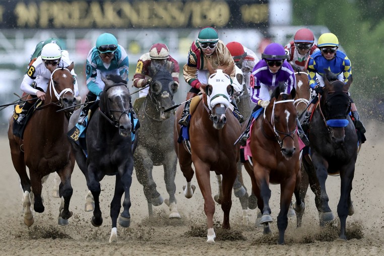 A tight group of racehorses and jockeys race toward the camera on a dirt track.
