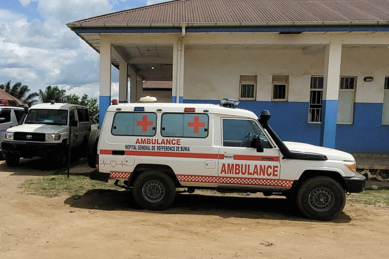 A white 4x4 with red lettering reading AMBULANCE parked on a dirt road outside a one-story concrete building.