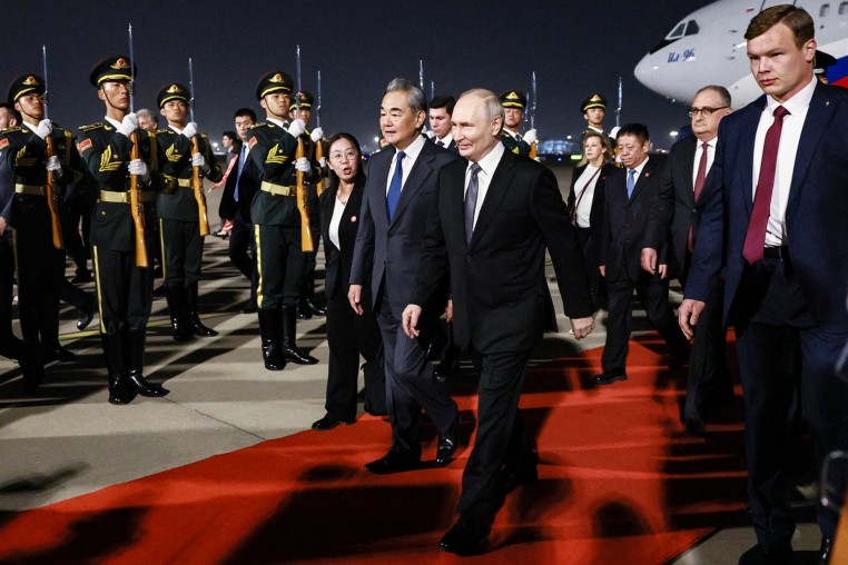 Vladimir Putin and Wang Yi walk along a red carpet extending from a plane parked in the background, with soldiers and other officials surrounding them