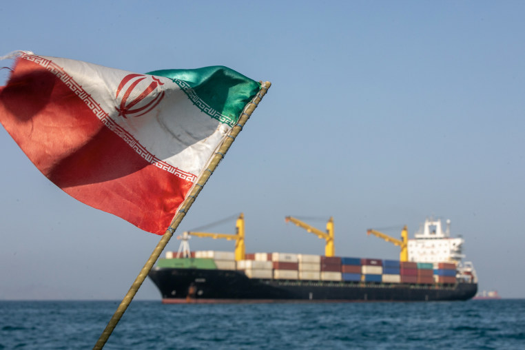An Iranian flag appears in the foreground with a cargo ship in the background.