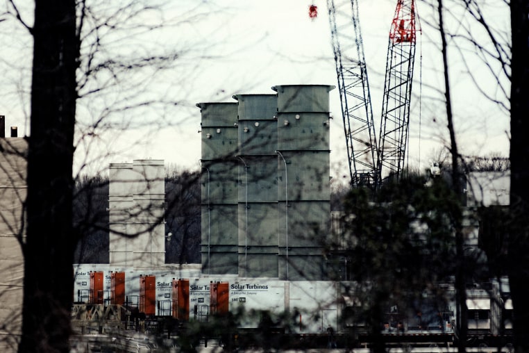A construction site with tall smokestacks in a wooded area under gray skies.