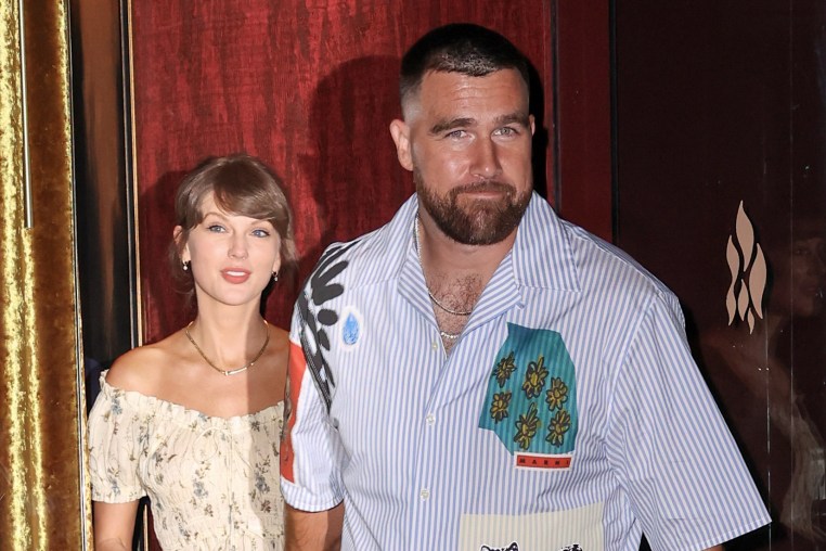 Travis Kelce, looking toward the camera, holds Taylor Swift's hand as they exit a building to the street.