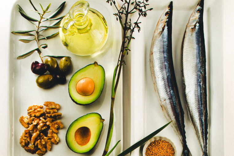 Still life of foods like olives, canola oil, mackerels, flaxseeds, avocado, and walnuts. 