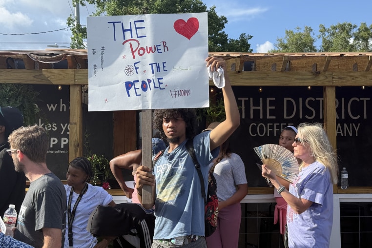 A young man holding a sign reading THE POWER OF THE PEOPLE at a small protest. 