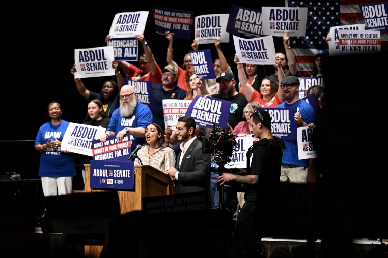 Abdul El-Sayed and Alexandria Ocasio-Cortez on stage during a campaign rally, surrounded by people holding ABDUL FOR SENATE signs.