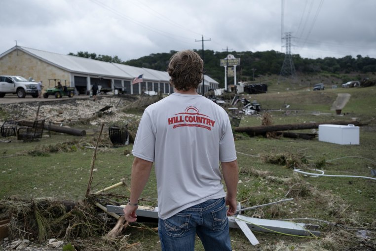 A man wearing a shirt that says HILL COUNTRY stands in front of flood damage strewn across a lawn.