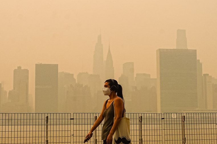 A woman wearing a facemask walks outside in an orange smoky haze past the New York City skyline