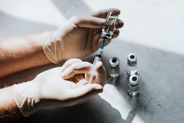 A hand wearing latex gloves hold a phial of clear liquid and a syringe. The medical professional uses the syringe to extract the medicine from the glass bottle. 