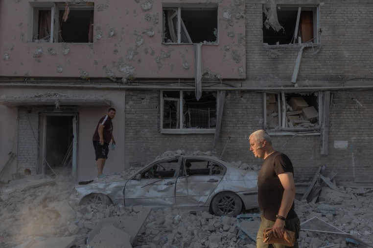 A man walks past while a man stands on a destroyed car following an air attack.