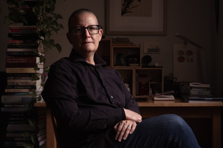 A middle-age white woman with a shaved head sits with crossed legs in front of her desk and large stacks of books.