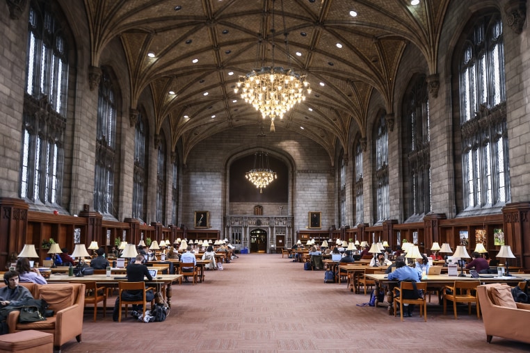 A large stone room with a vaulted ceiling and chandelier and long rows of wooden desks.