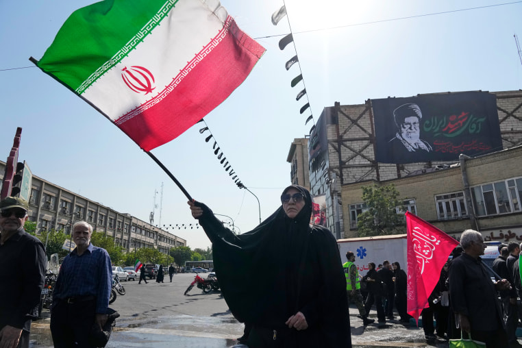 Image: A woman waves an Iranian flag. A banner showing a portrait of the late Supreme Leader Ayatollah Ali Khamenei is seen hanging on a building wall in the background.