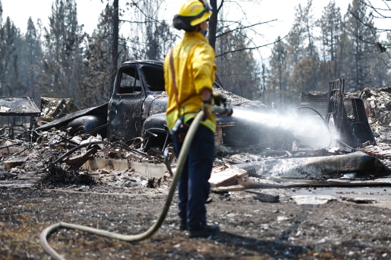 A firefighter sprays water from a hose onto debris near a charred car outside