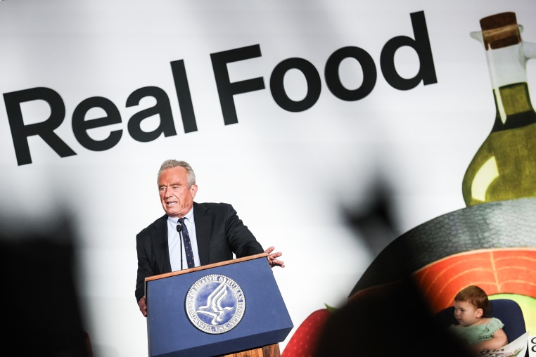 Robert F. Kennedy Jr speaks at a podium onstage, in front of a white backdrop with black text above him that reads "Real Food"