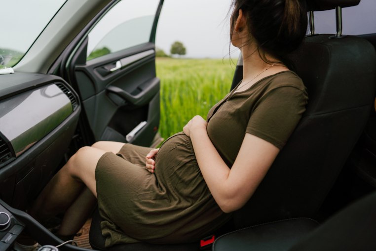 A pregnant woman sitting in a car in the passenger seat looks out at the view, as seen from the view from the driver's seat