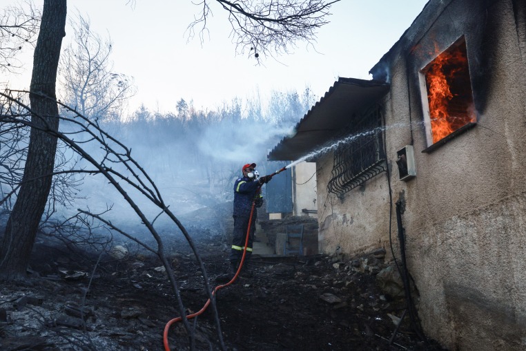 A firefighter sprays a hose into the window of a house on fire, in a charred forest environment