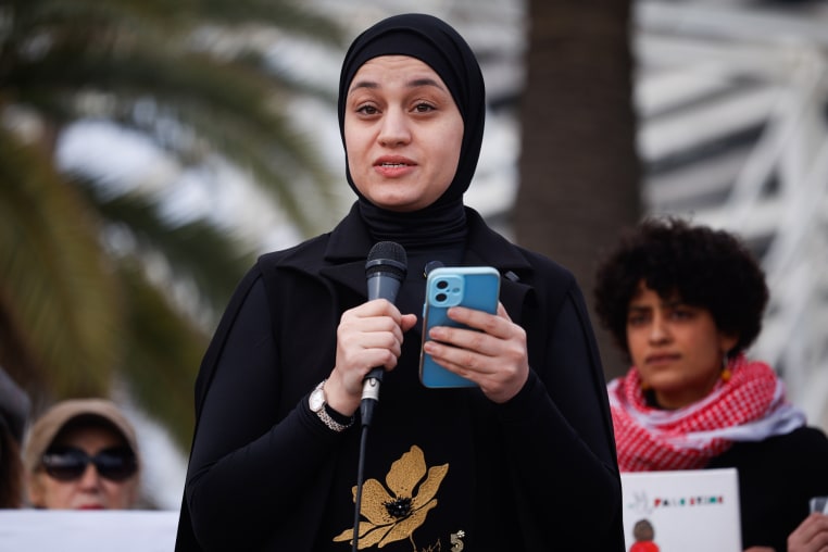 Wissam Hamada speaks into a microphone while standing and hold a cellphone in the other hand