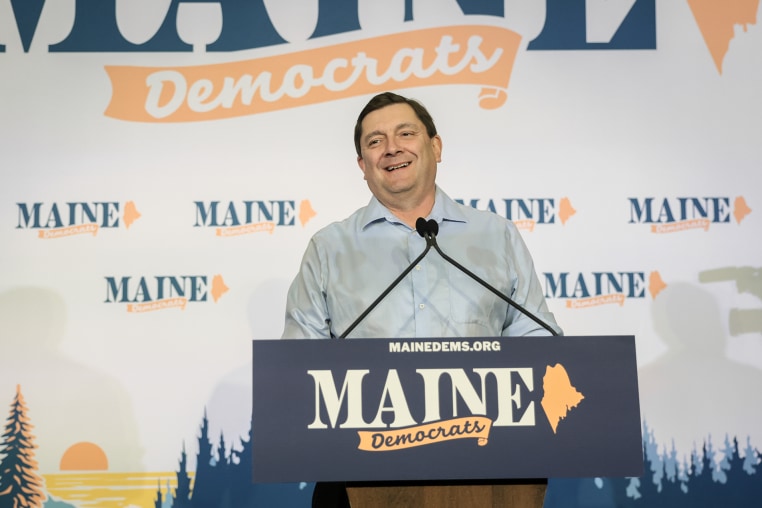 Troy Jackson smiles while standing at a lectern with a "MAINE DEMOCRATS" logo on it.