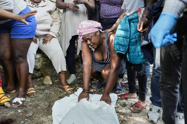 A crying woman leans over a body covered in white tarp outside, surrounded by others