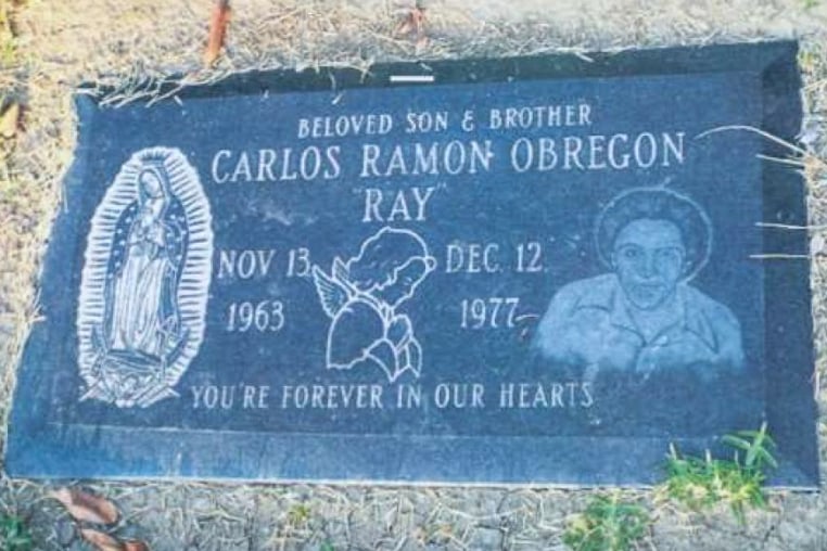 Carlos Ramon Obregon's headstone, flat on the ground, showing an engraving of Mother Mary on the left, a praying angel in the center, and Carlos on the right. The full headstone reads "Beloved Son & Brother, Carlos Ramon Obregon, "Ray", Nov. 13, 1963 - Dec. 12, 1977, You're forever in our hearts"