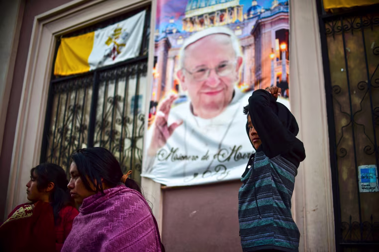 People stand on a street with a portrait in the background of the Pope Francis.